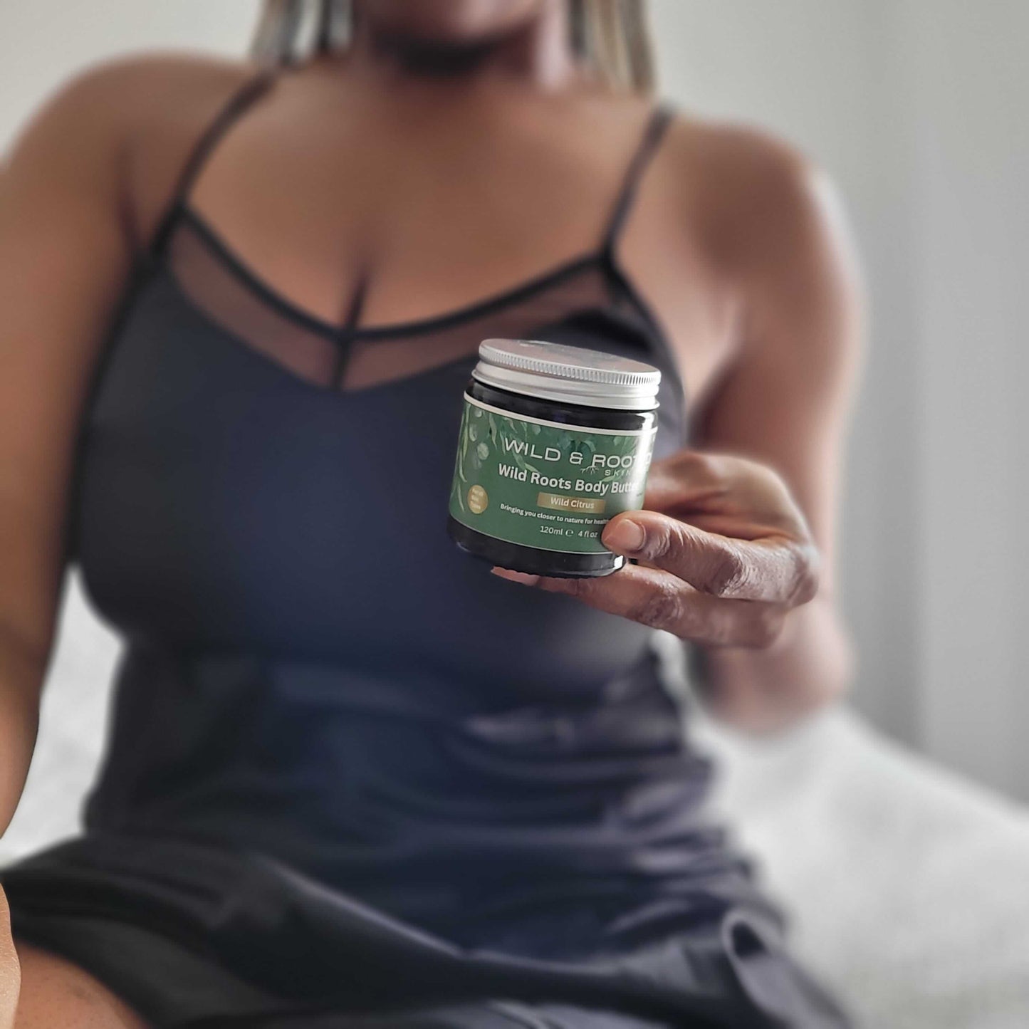 A woman sitting on a bed in a nightdress holding a jar of Wild Roots body butter